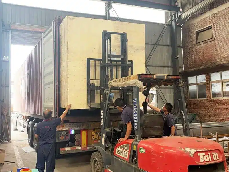 Workers using a forklift to load a large wooden crate containing bumper car equipment into a shipping container at the factory.