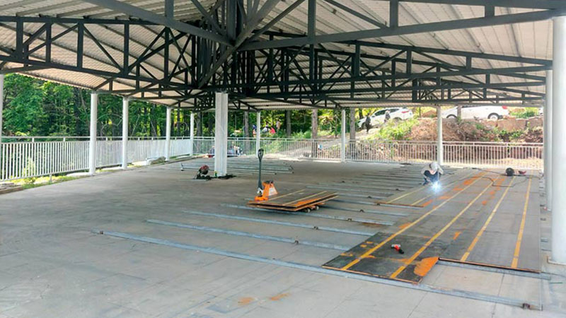 Workers welding and assembling modular conductive floor panels for ground grid bumper cars inside a sheltered pavilion.