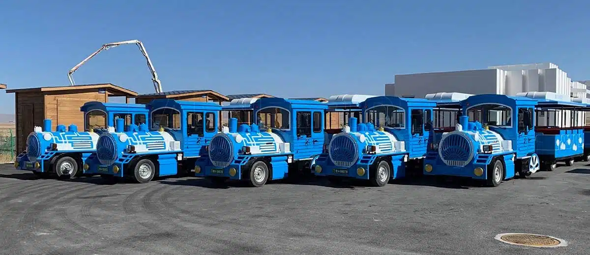 أسطول كبير من قطارات الملاهي الزرقاء المجمعة حديثًا بدون مجنزرات مصطفة في الهواء الطلق. توفر شركة كارني رايدس (Carnee Rides) بصفتها موردًا رائدًا في الصين طلبيات بالجملة من هذه القطارات الكهربائية شديدة التحمل لمدن الملاهي الكبيرة.