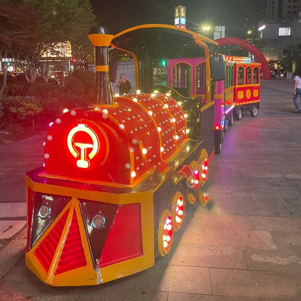 A red outdoor classic trackless sightseeing train adorned with bright LED bulbs, operating safely during the evening in a commercial plaza.