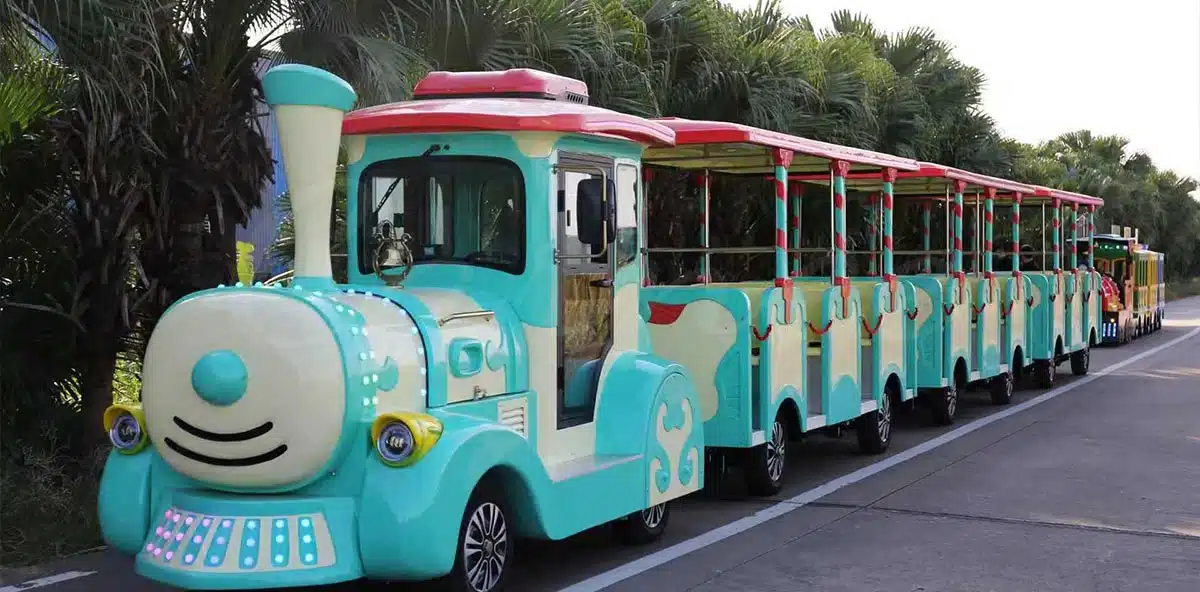 A light blue and white kiddie trackless tourist road train carrying passengers along a paved outdoor pathway in a scenic amusement park environment.