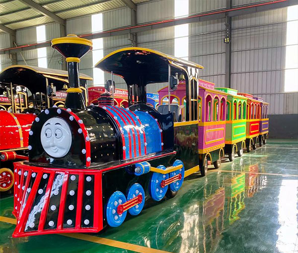 A classic blue and red electric trackless train ride with yellow trim, inspired by Thomas style, parked inside a manufacturing factory workshop.