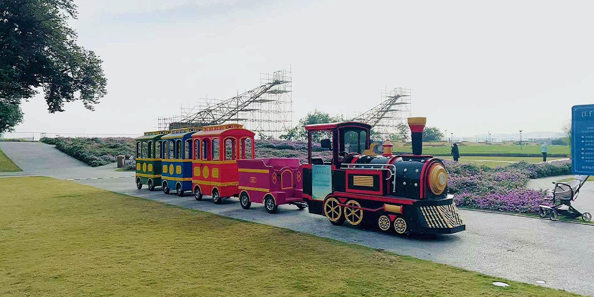 Classic black and red vintage steam-style trackless train with colorful carriages cruising along a curved pathway through a green park and flower field.